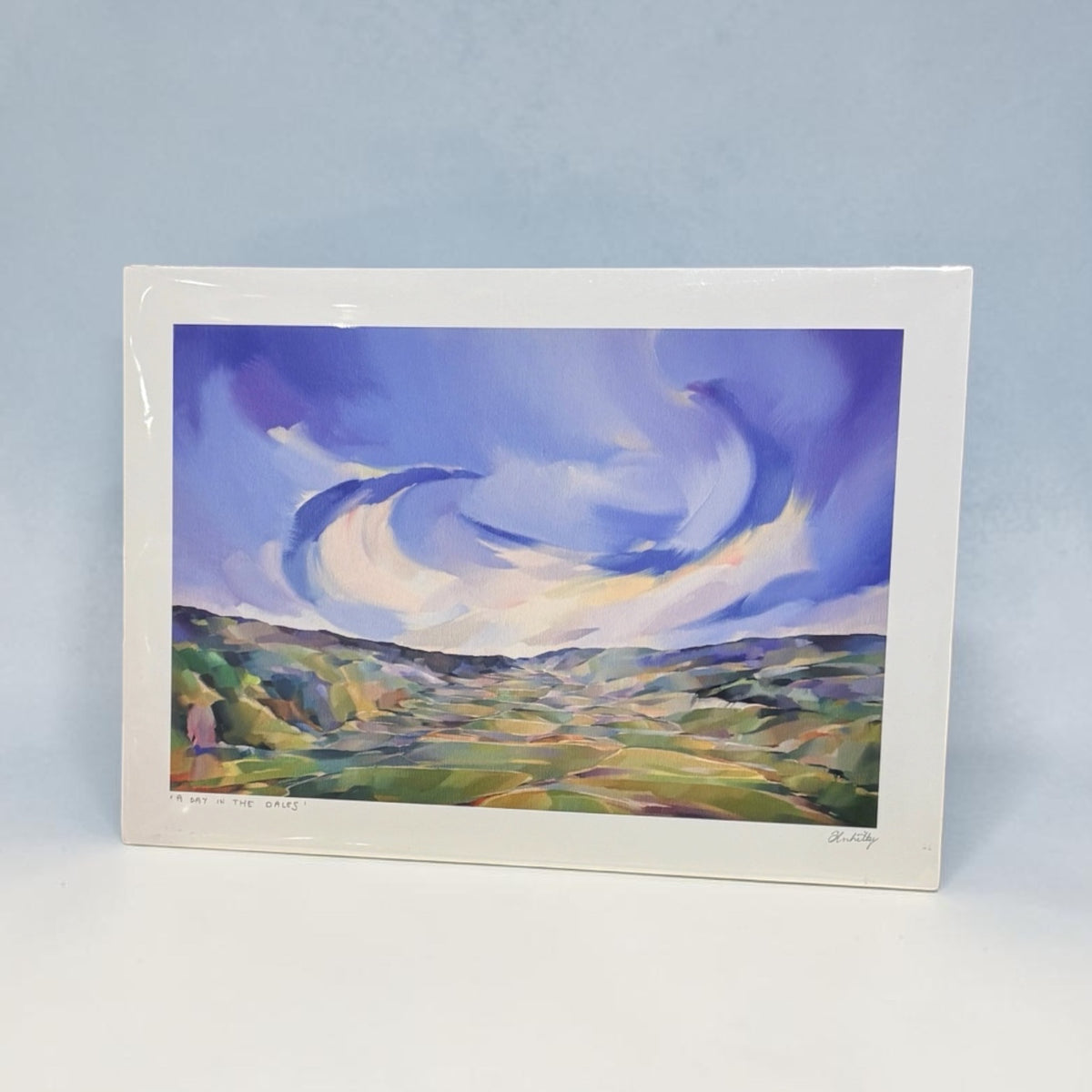 A framed landscape art print showing a stylized blue sky and green valley, photographed against a light blue background.