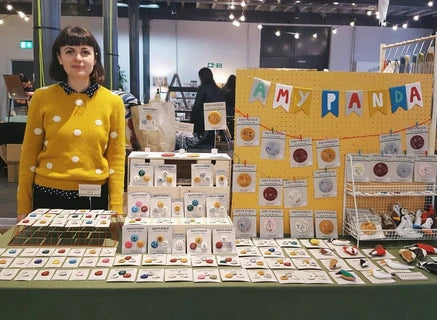 A woman stands behind a craft display table at an indoor market or fair. The table is covered with handmade items, including small packaged pieces, cards, and a yellow pegboard sign that reads 'AMY PANDA.