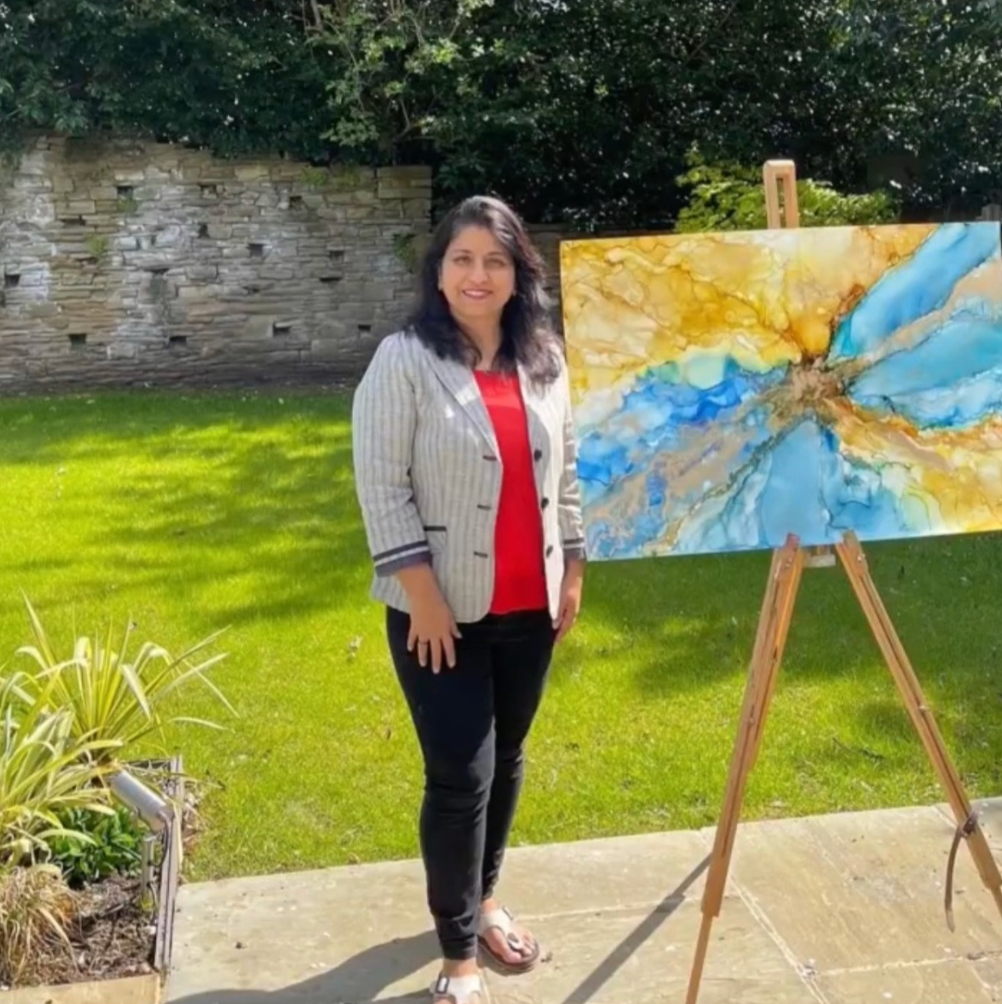 A woman stands outdoors on a patio next to an easel holding a colorful abstract painting with blue, gold, and white tones. She is wearing a gray pinstripe blazer over a red top and black pants, with a green lawn and stone wall in the background.