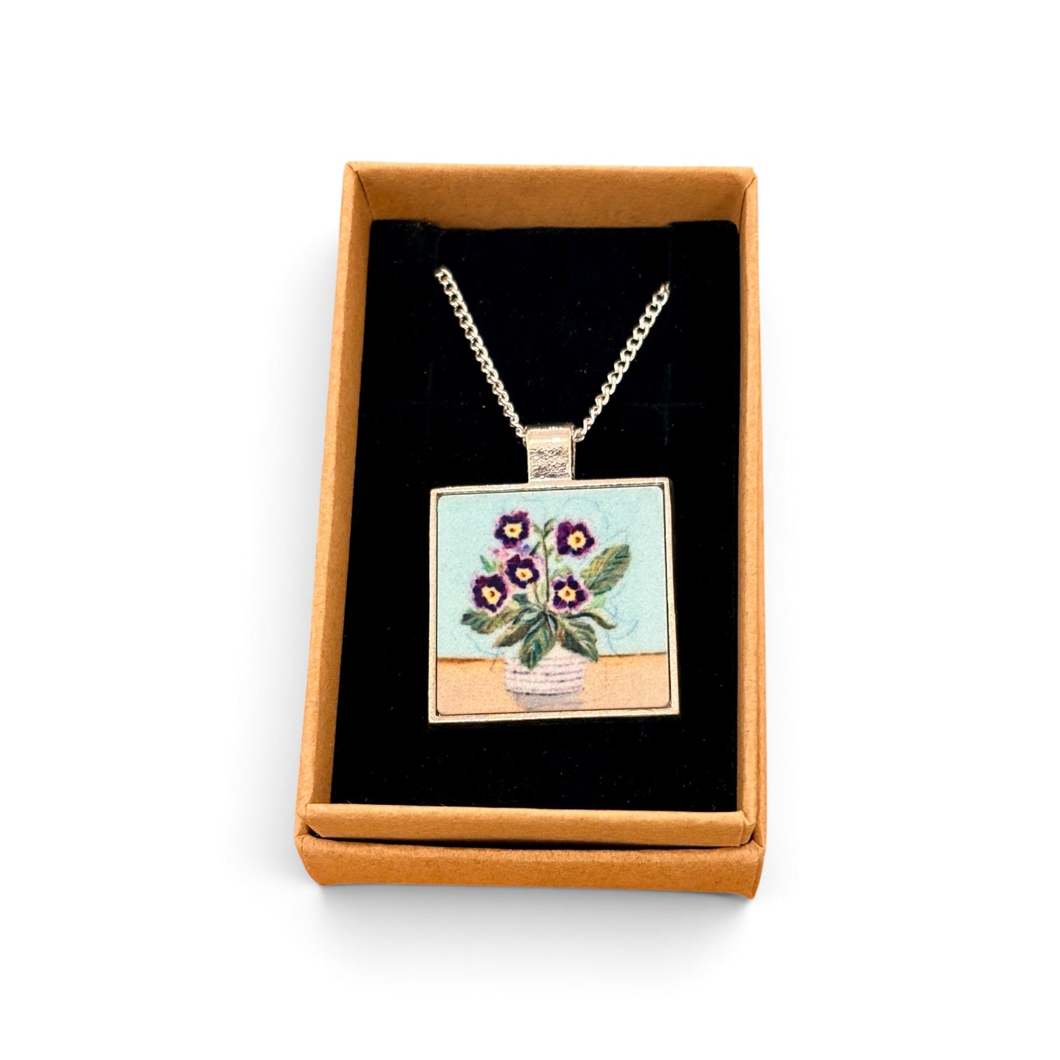 A square pendant necklace displayed inside an open tan gift box with a black insert. The pendant shows a floral bouquet in a vase with purple flowers and green leaves on a light blue and beige background, suspended from a silver-toned chain.