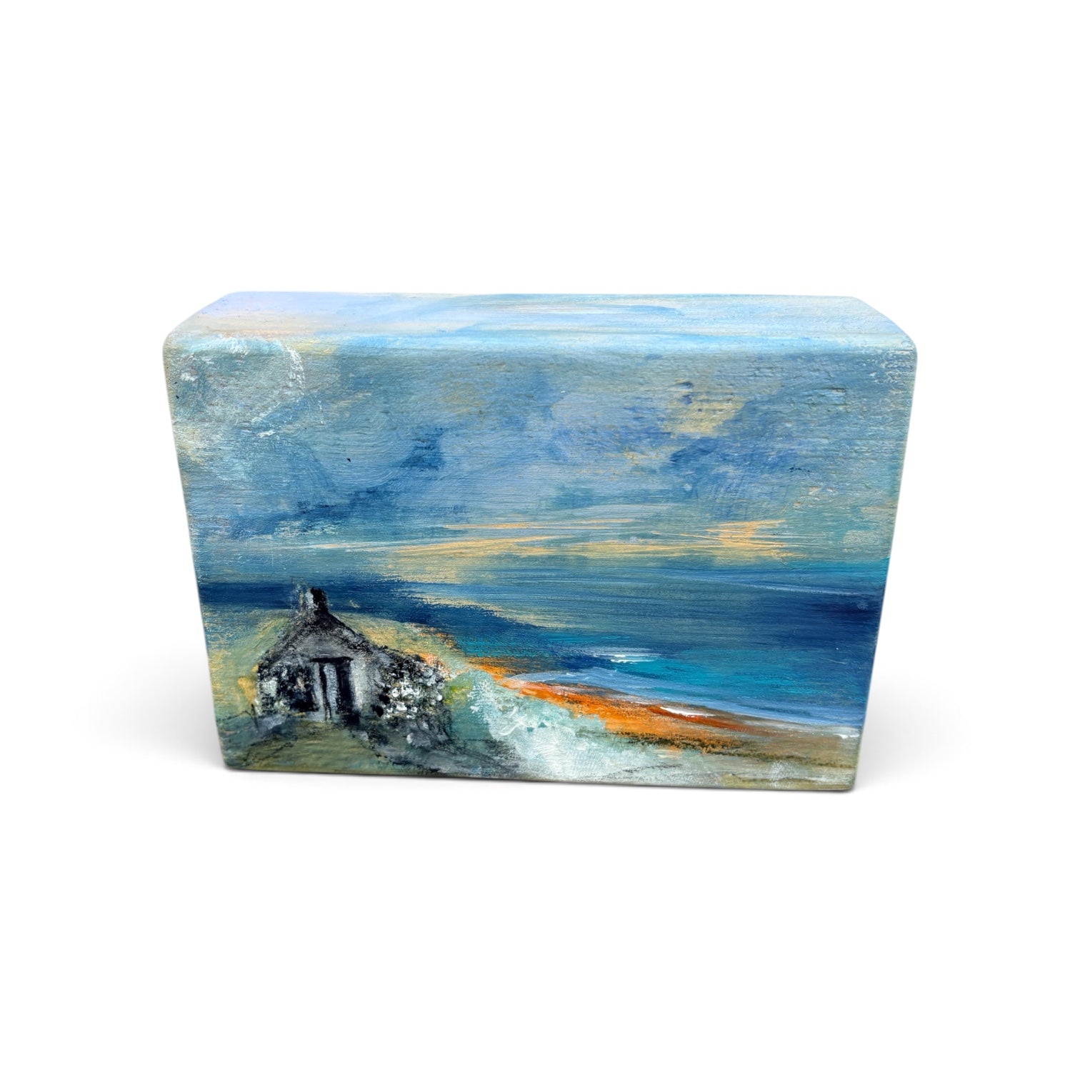 A rectangular wooden plaque or box featuring an impressionistic painting of a small cottage by the sea, with blue and gray sky, ocean waves, and a sandy shore, shown on a plain white background.