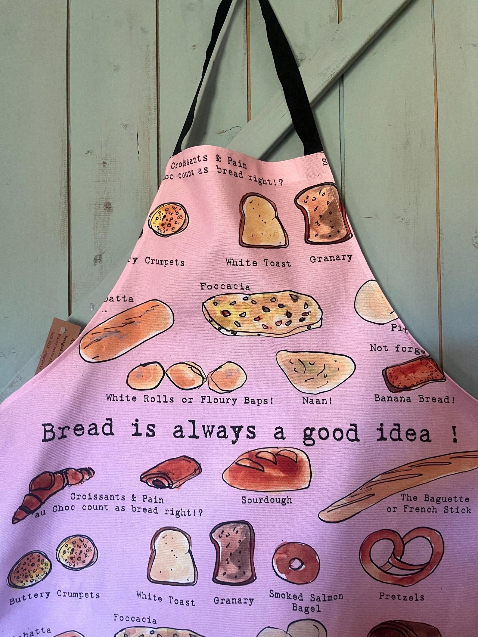 A pink kitchen apron with black straps, featuring a playful print of various breads, croissants, and pastries with humorous labels like &