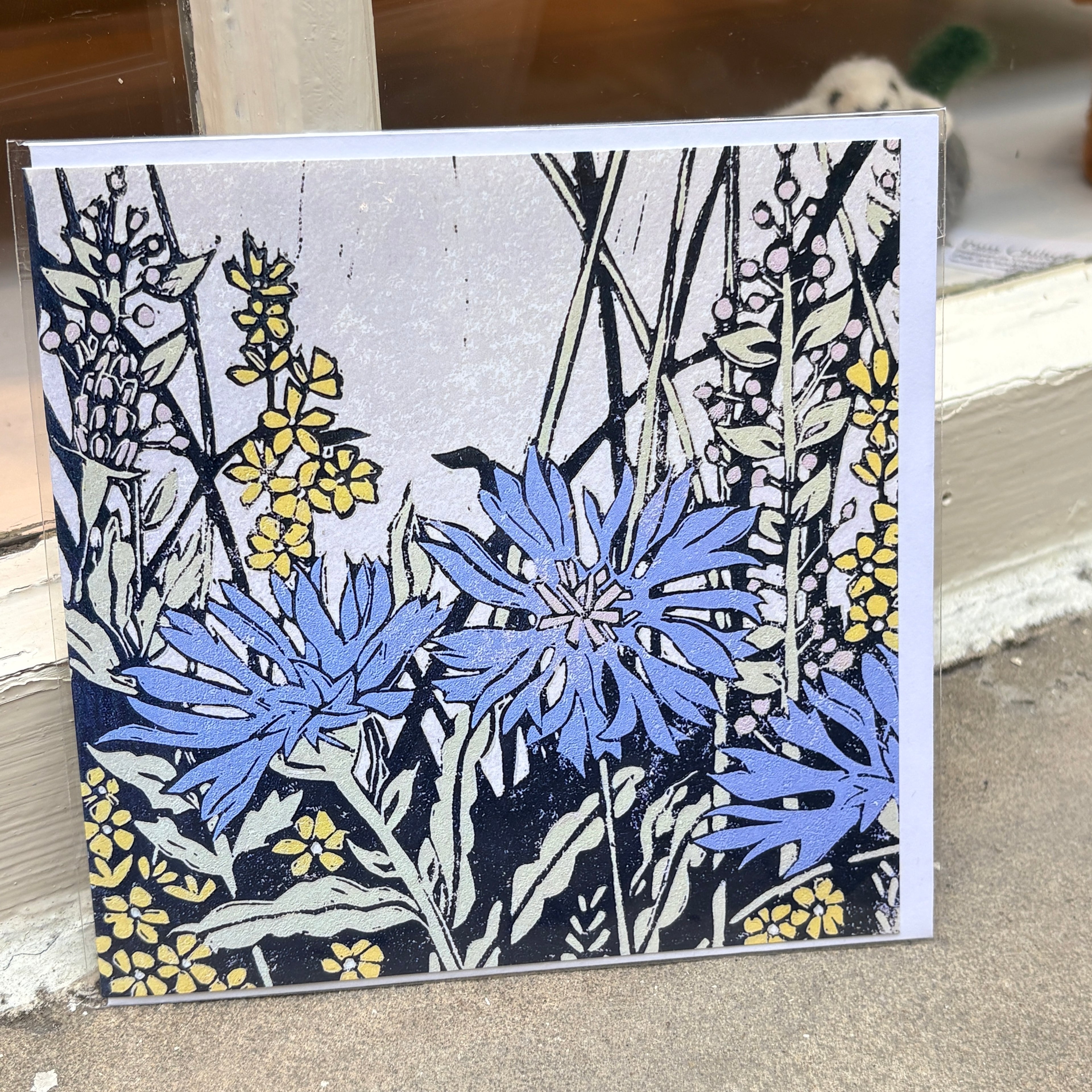 A square art print or card featuring a botanical illustration of blue cornflowers and yellow flowers with black linework on a light gray background, photographed leaning against a doorway or shelf.