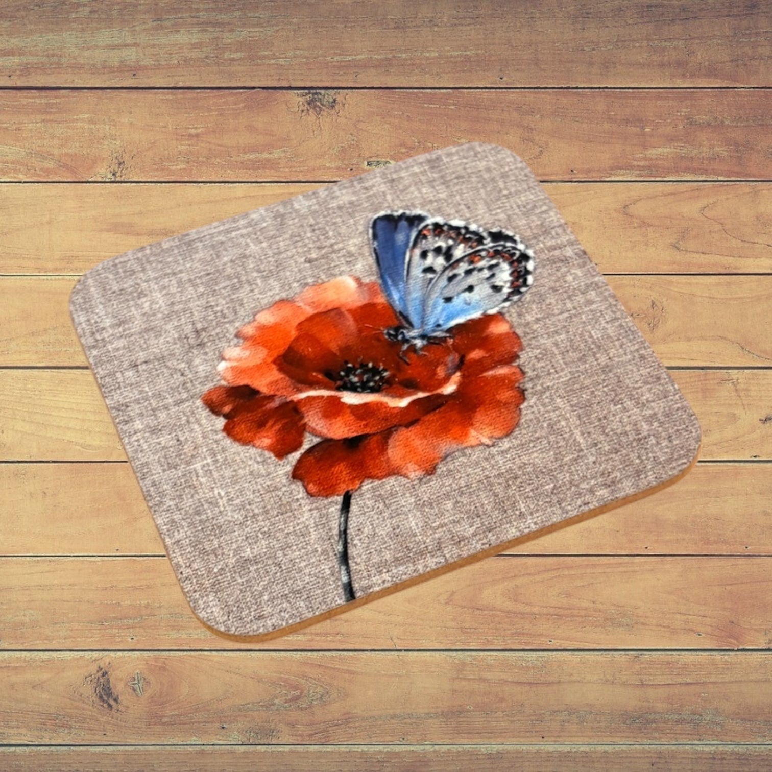A square cork placemat with rounded corners featuring a printed design of a blue butterfly resting on a red-orange poppy flower, displayed on a rustic wooden plank surface.