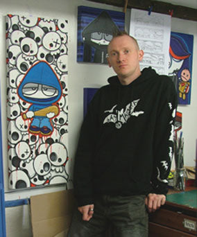 A man wearing a black graphic hoodie stands in front of a wall covered with art prints and sketches, next to a large colorful cartoon print featuring a blue hooded figure and white skulls.
