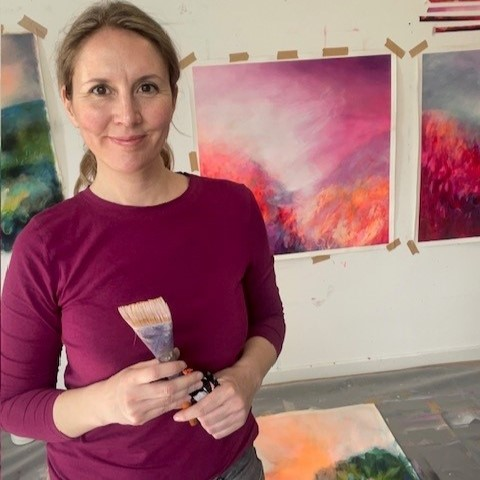 A woman with blonde hair tied back, wearing a magenta long-sleeve shirt, holds a paint brush in front of a white wall decorated with colorful abstract paintings.