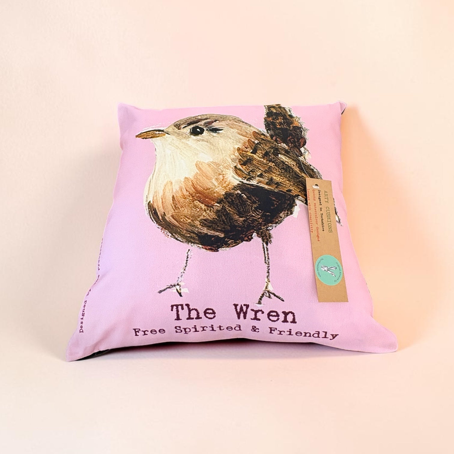 A square pink pillow featuring a painted bird illustration labeled &