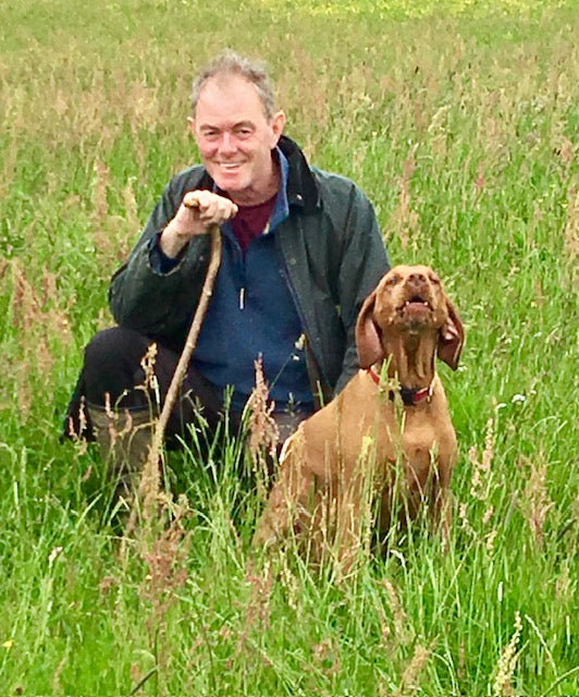 A man kneeling in a grassy field with a brown dog, holding a rope leash. The man is wearing a dark green jacket over a blue shirt, and the dog has a collar.