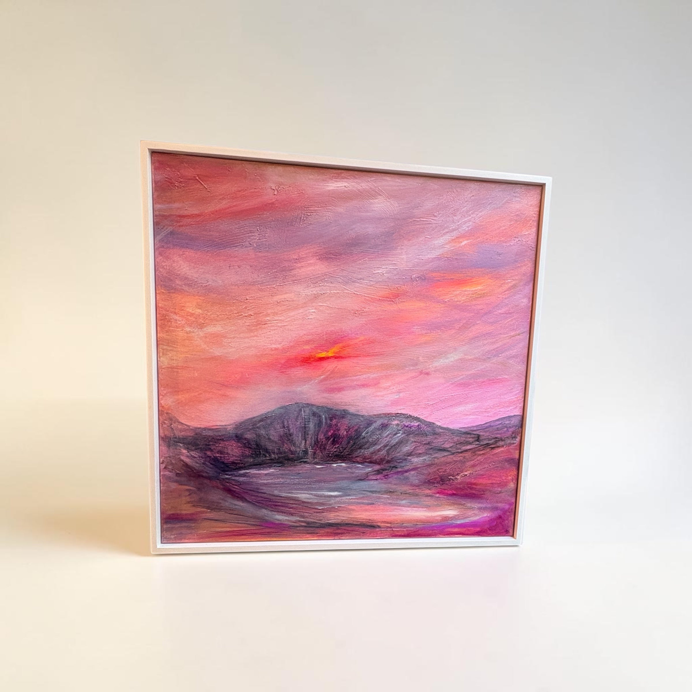 A square framed painting of a mountain landscape at sunset, featuring vibrant pink, purple, and orange hues, displayed on a clean white surface against a light gray background.