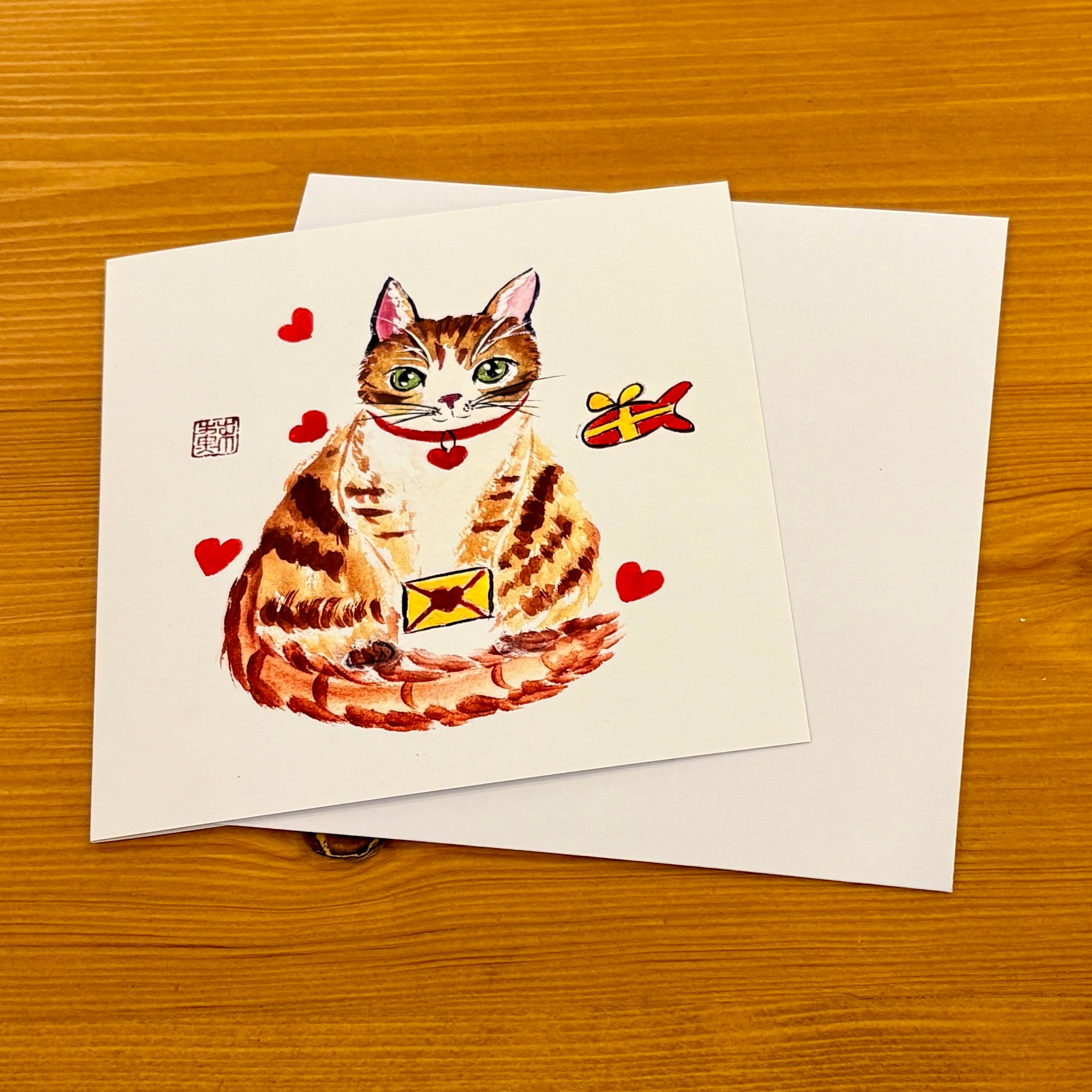 A square white greeting card featuring a watercolor-style orange tabby cat with red hearts and a small yellow envelope, shown on a wooden table with an additional white card behind it.