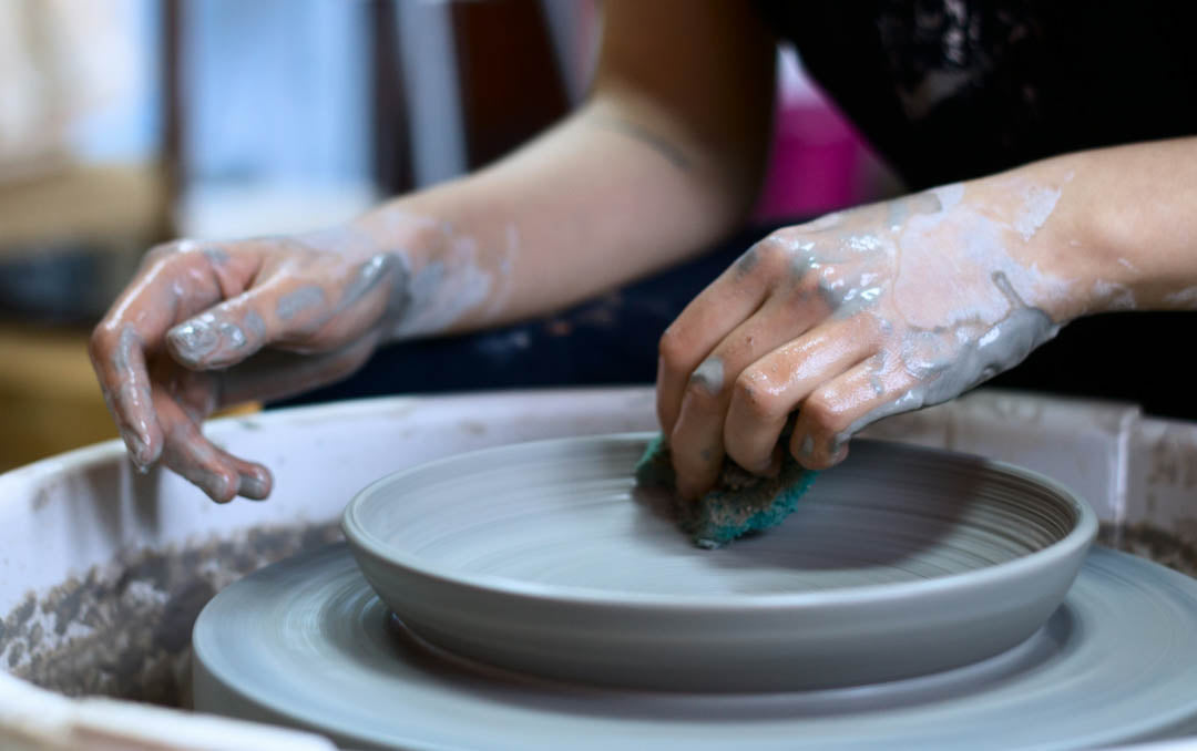 Image depicts a pair of woman's hands at a pottery wheel creating a bowl.