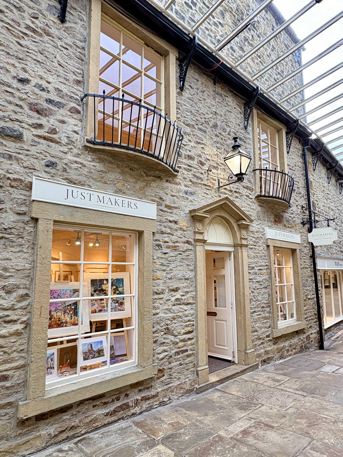 A stone shopfront building with a sign reading JUST MAKERS above a large window display, set in a covered outdoor alley with a glass roof and stone pavement.