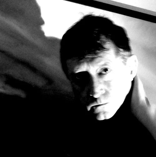 Black and white close-up portrait of a man looking directly at the camera, with dramatic high-contrast lighting and shadows.