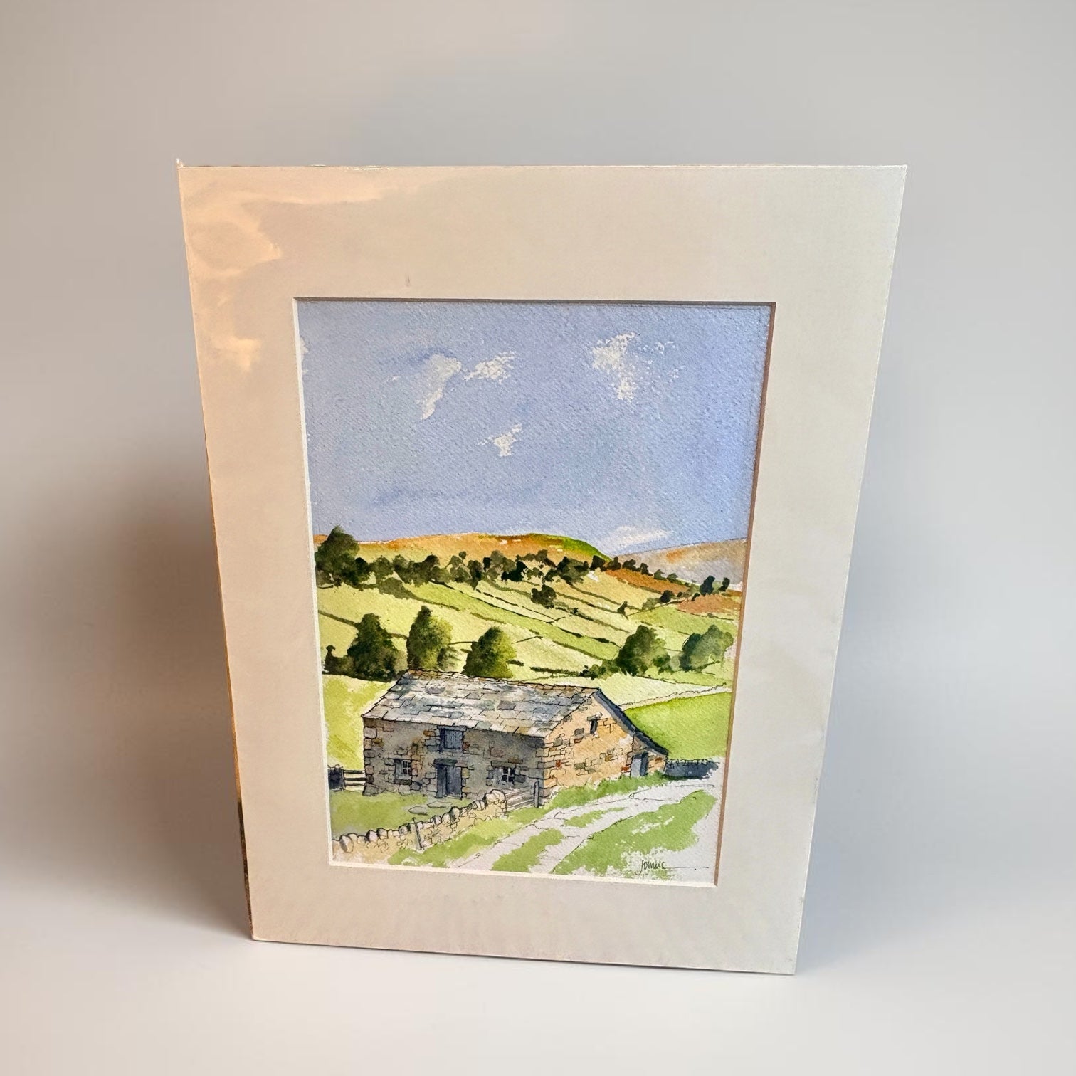 A framed watercolor landscape print showing a stone cottage on a grassy hillside under a blue sky, photographed against a neutral light gray background.