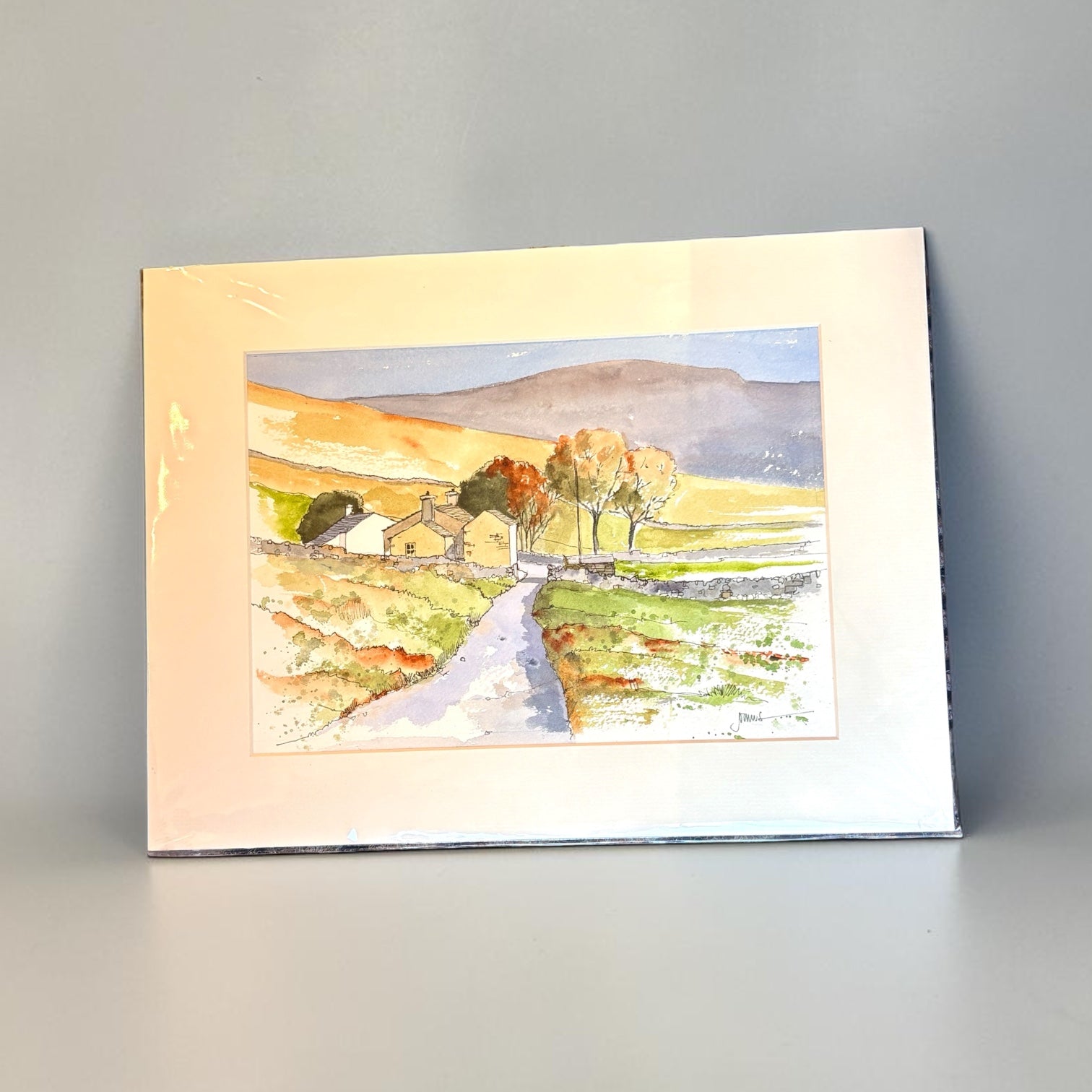 A watercolor landscape art print depicting a rural scene with a winding path, small cottages, trees, and rolling hills, shown with a wide cream mount against a neutral gray background.