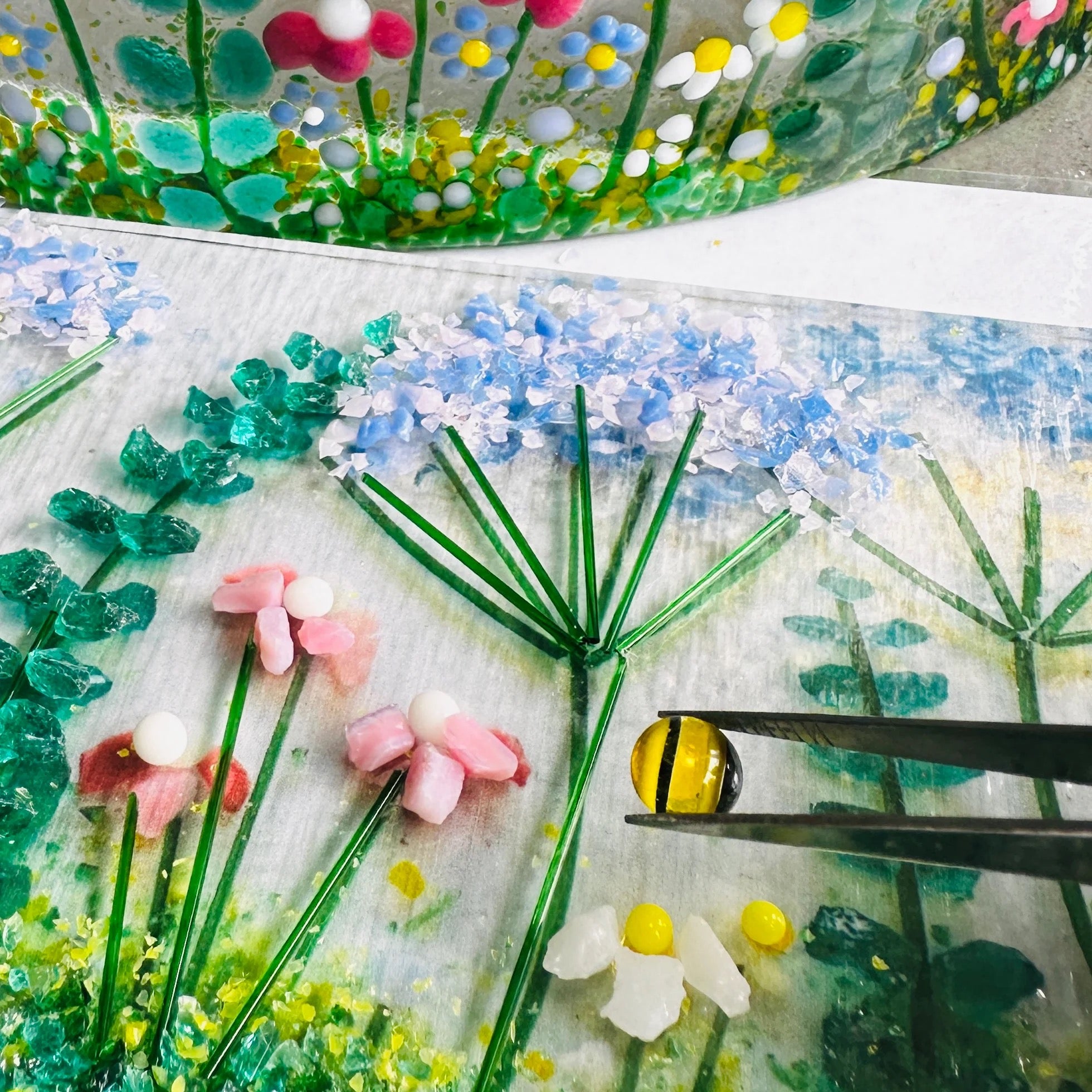 Close-up of a handcrafted glass garden scene with flowers and a bee, using tweezers.