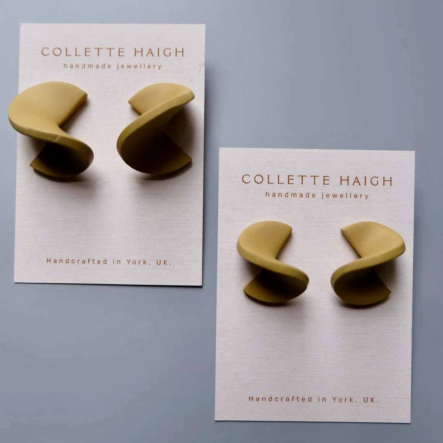 Two pairs of Collette Haigh handmade spiral stud earrings in a muted olive-gold tone, mounted on white display cards against a light gray background.