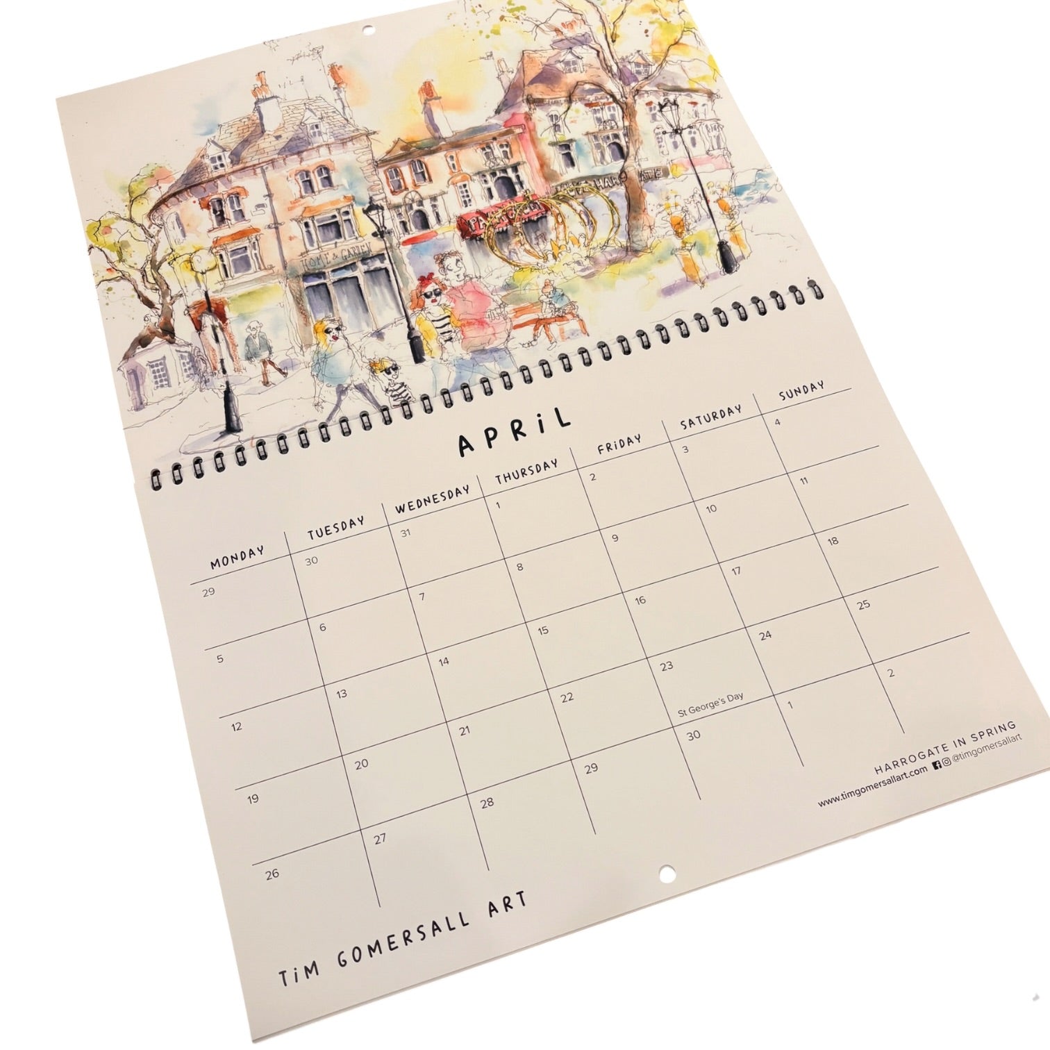 A spiral-bound April desk calendar page from Tim Gomersall Art, featuring a colorful watercolor illustration of a street scene with houses, trees, and people above a printed calendar grid.