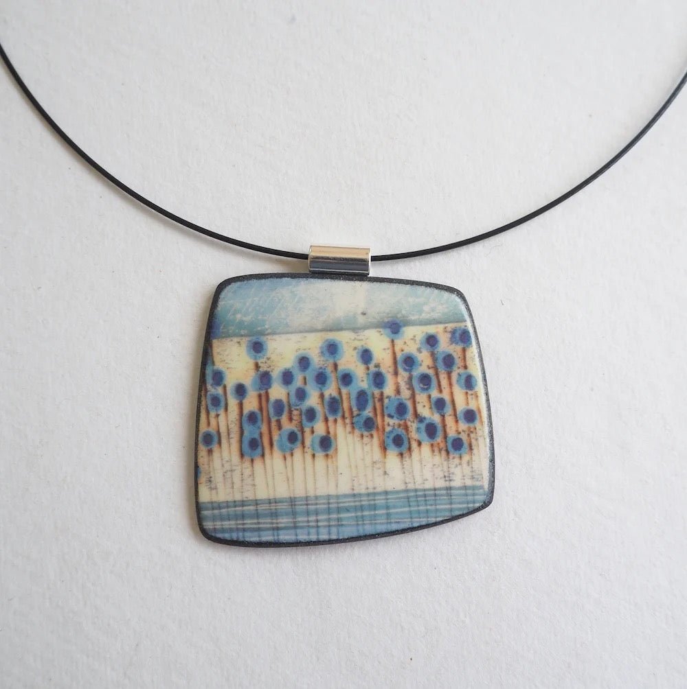 Square pendant with blue and beige pattern on a black cord against a light gray background
