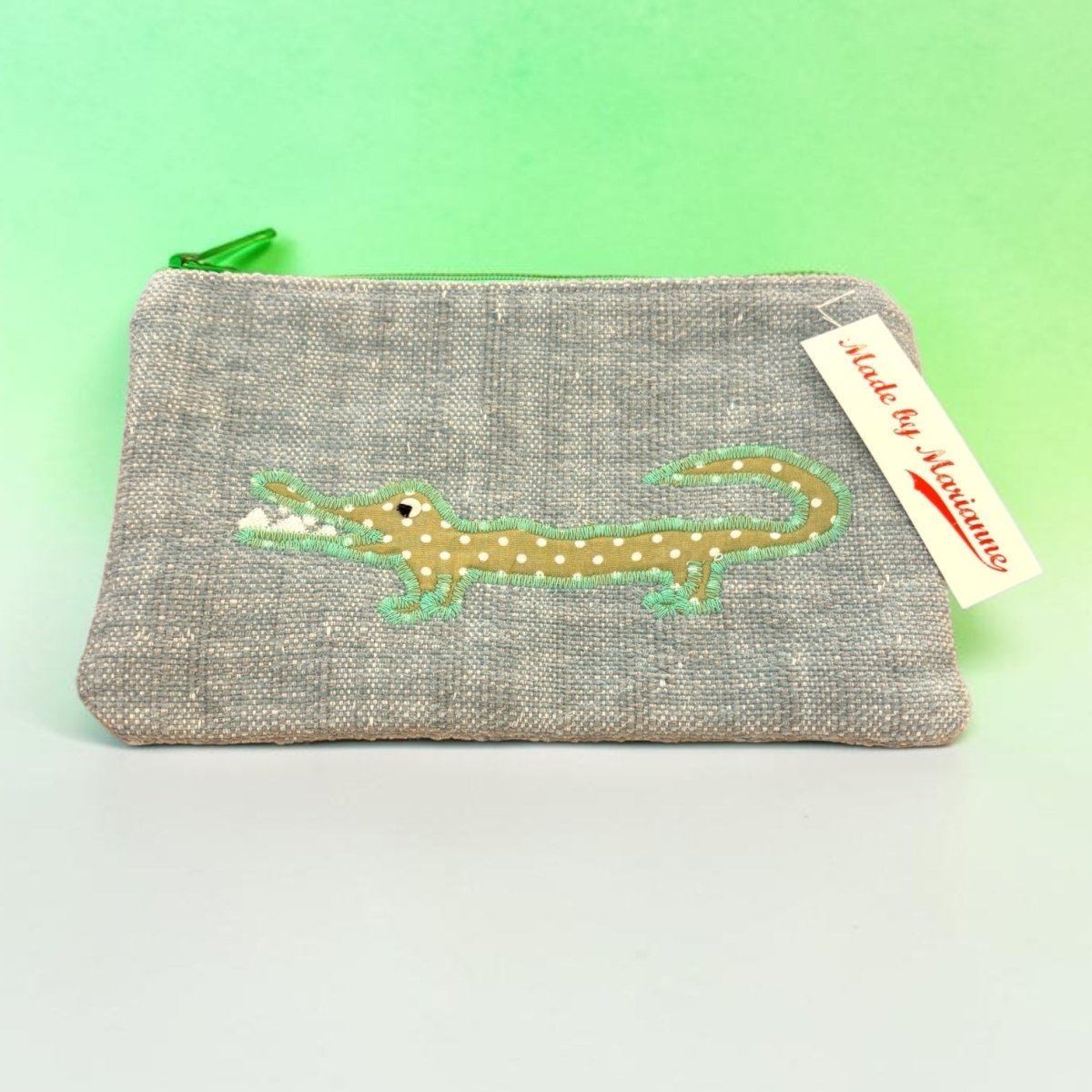 A gray woven fabric zippered pouch featuring an embroidered alligator design in tan with white spots and green outlines. A green zipper pull is visible on the left, and a white &