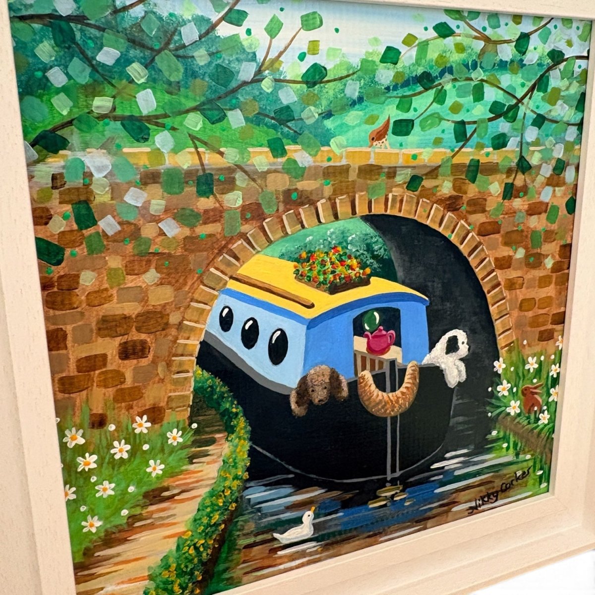 A colorful framed painting depicting a blue and yellow canal house on water under a brick bridge, surrounded by green foliage and small white flowers. The artwork is shown inside a light wood frame.
