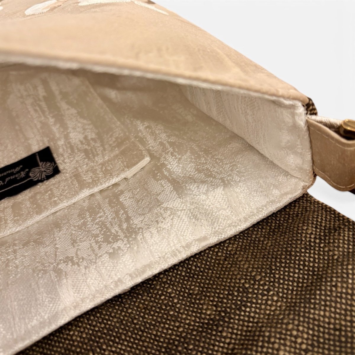 Close-up of a beige fabric handbag showing the interior lining and a black brand label, with a brown woven exterior visible at the bottom.