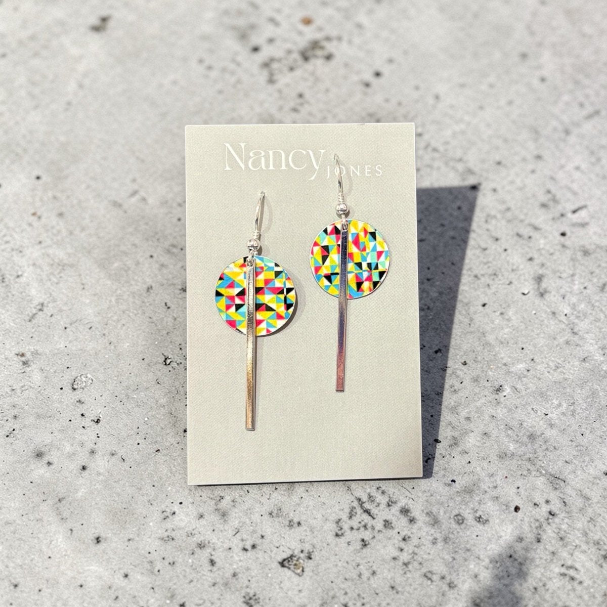 Colorful geometric earrings on a Nancy James card against a textured gray background
