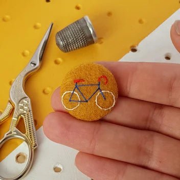 A hand holds a small round felt pin featuring an embroidered bicycle design in blue, red, and white. In the background, gold scissors and a metal thimble rest on a yellow and white pegboard surface.