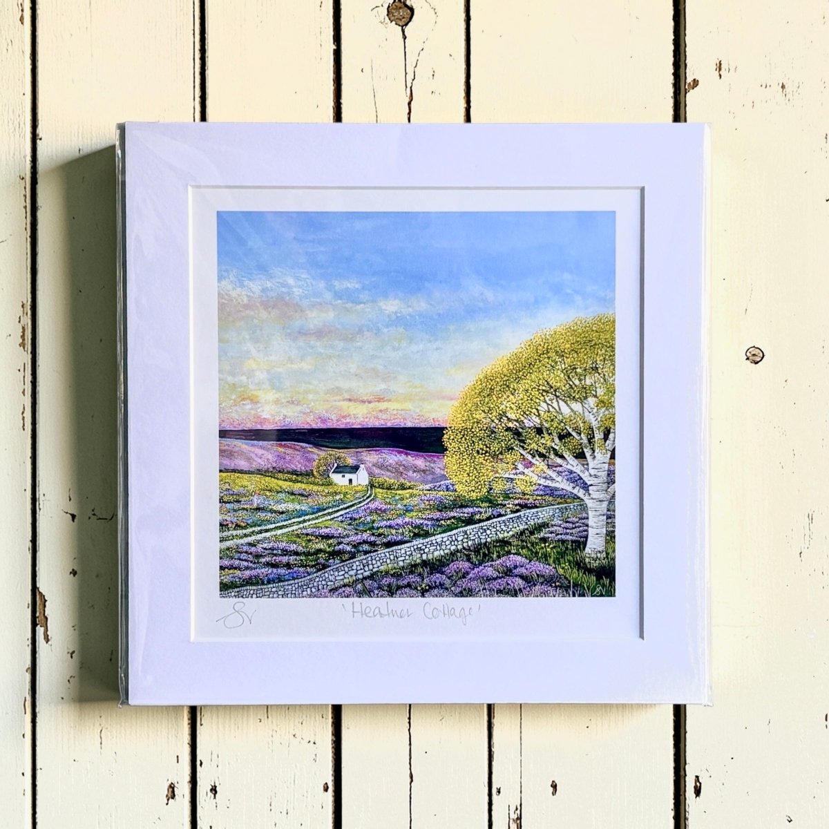 Framed artwork of a landscape with a house, tree, and lavender field on a wooden surface.