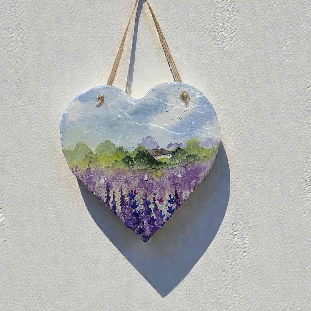 Heart-shaped painting of a landscape with lavender fields and a house, hanging on a textured white wall.