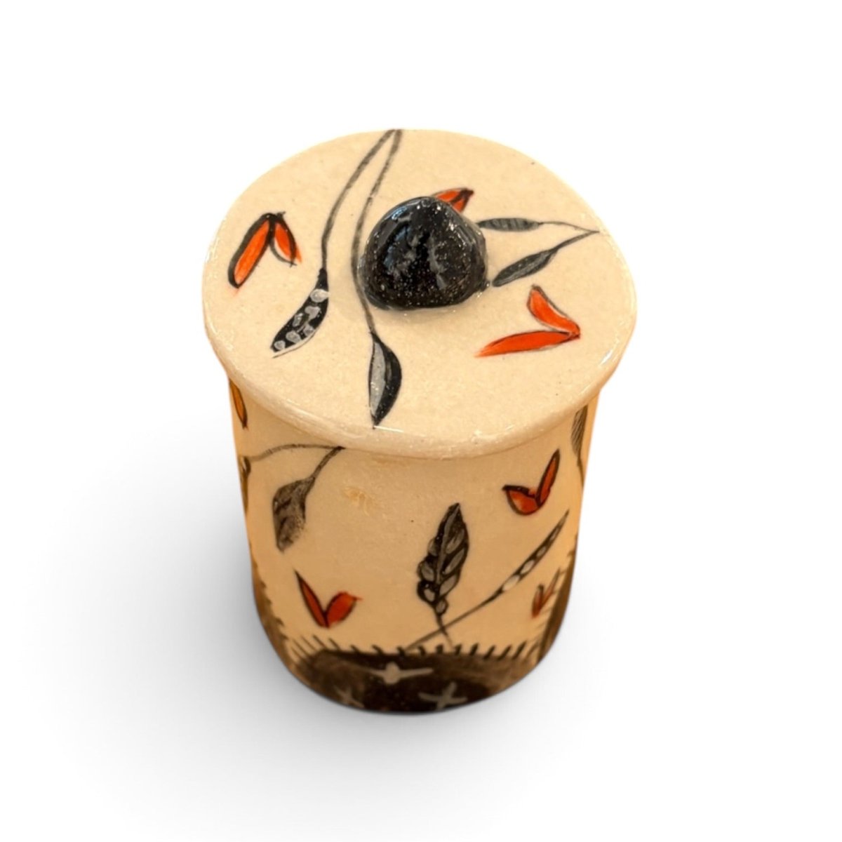 A small cylindrical ceramic container with a lid, decorated with hand-painted black and orange botanical motifs on a warm beige background, set against a plain white studio background.