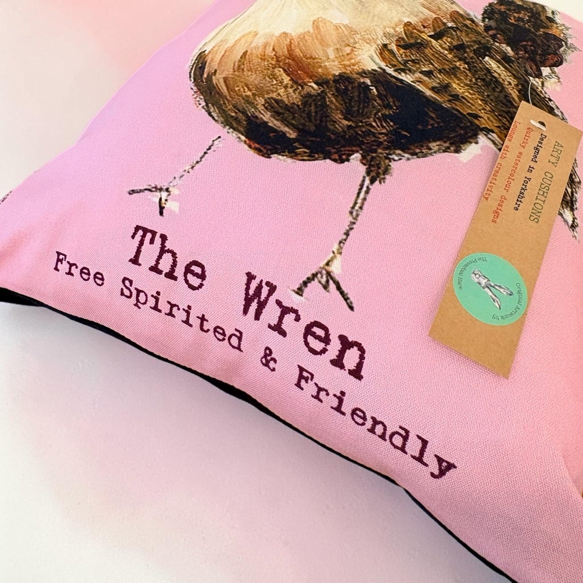 A pink decorative pillow featuring a painted wren illustration and the text &