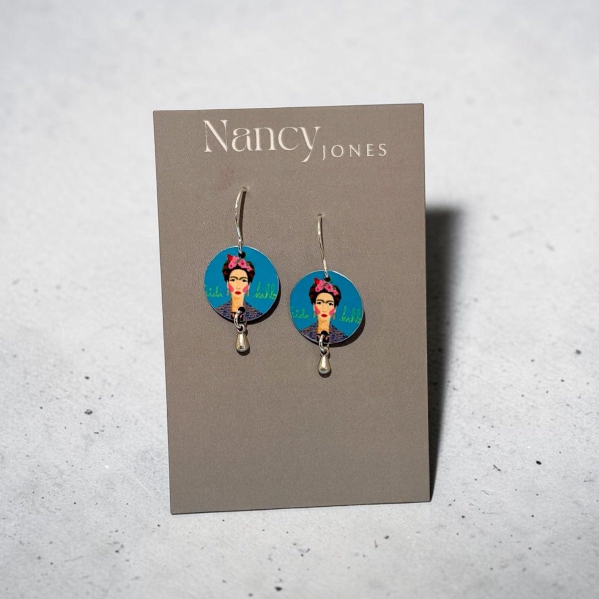 A pair of Nancy Jones dangle earrings displayed on a gray card. The earrings feature circular blue and pink graphic charms with a portrait illustration, suspended from silver hooks against a light gray textured background.