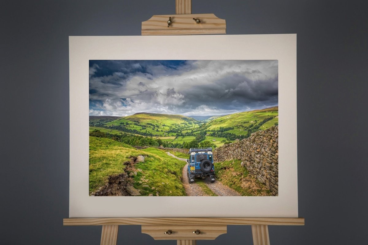 Framed landscape print of a rural scene with a tractor on an easel against a dark background