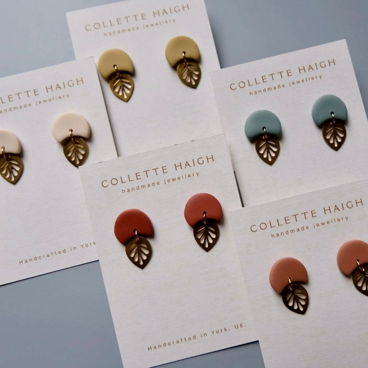 Six pairs of Collette Haigh handmade earrings displayed on their white backing cards, featuring various earthy tones including terracotta, dusty rose, sage green, cream, and light tan, with bronze leaf-shaped metal accents.