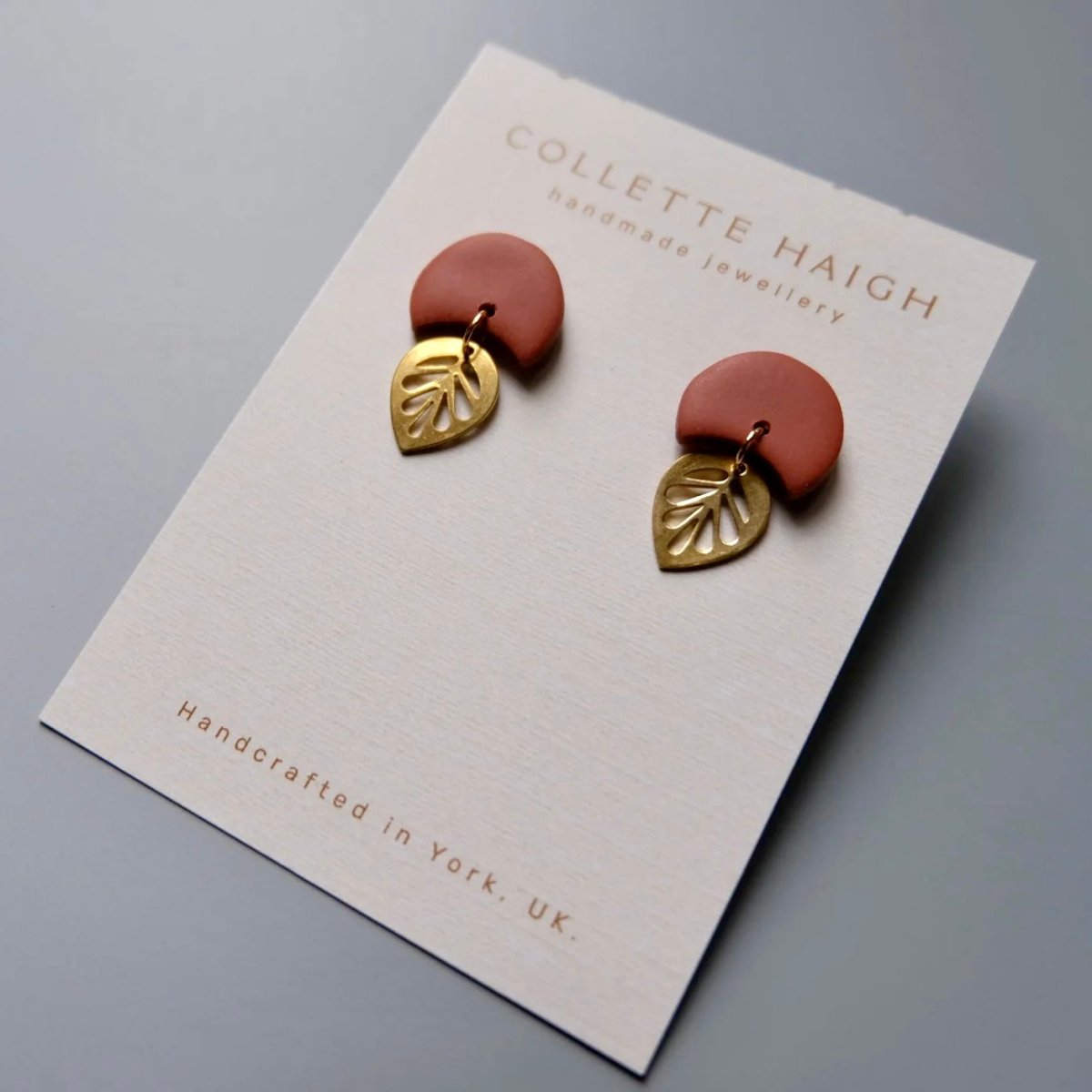 A pair of Collette Haigh handmade earrings displayed on a branded white card. Each earring has a terracotta-colored semi-circular top piece with a gold-toned leaf-shaped drop below, photographed on a smooth gray surface.