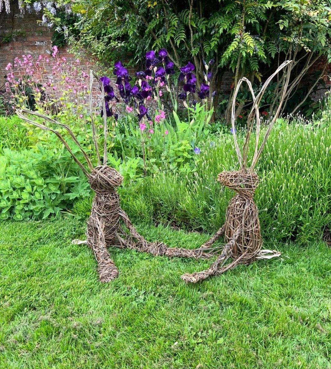 Two willow sculptures of a couple holding hands in a garden with flowers and greenery.