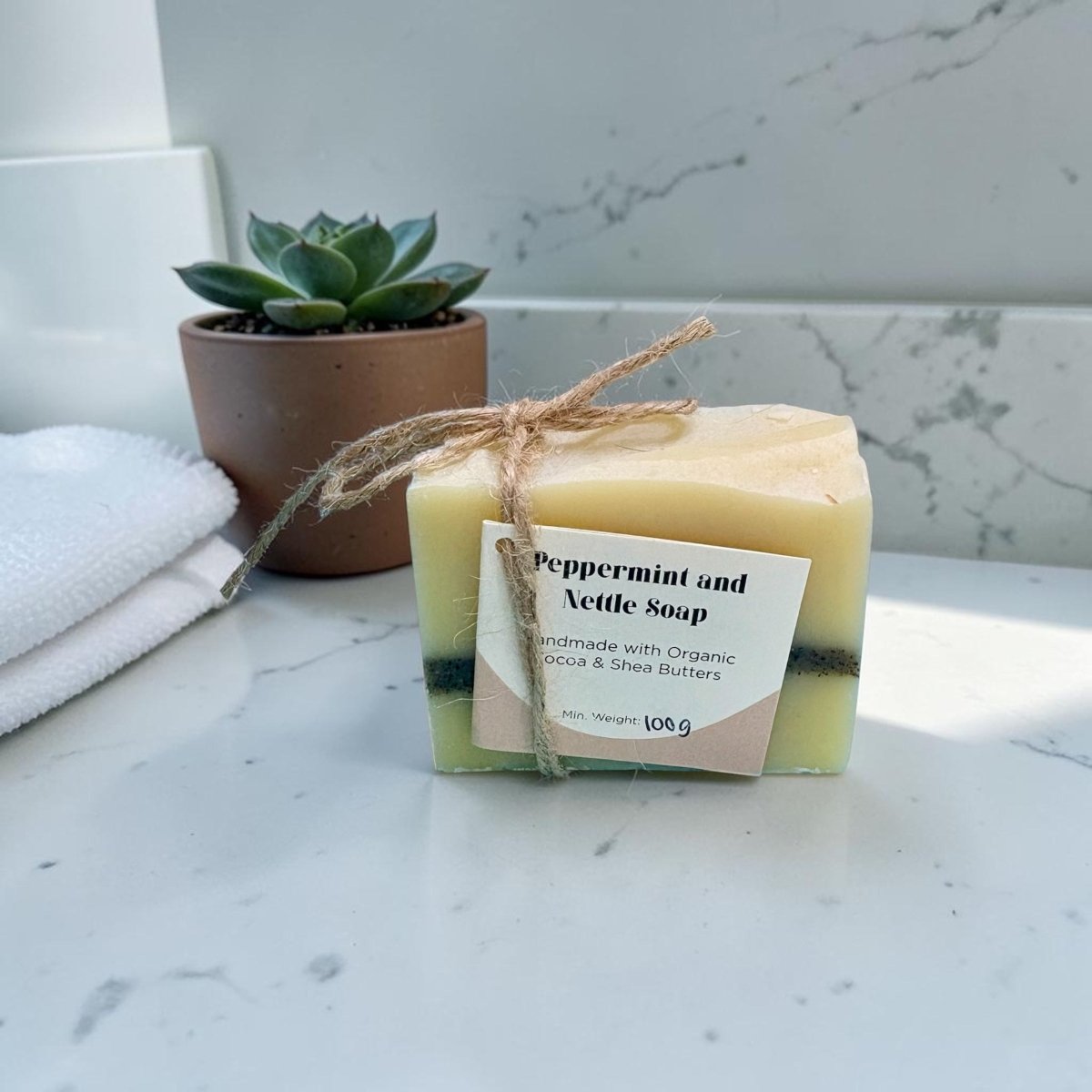 Bar of peppermint and nettle soap with a label on a marble surface, next to a potted plant and towel.
