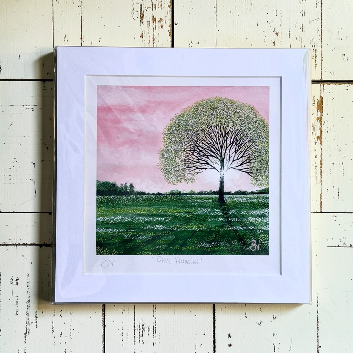 Framed artwork of a tree in a field with a pink sky on a wooden surface