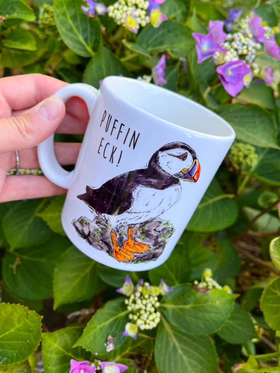 A hand holds a white ceramic mug featuring a black and white puffin illustration with orange accents and the text &