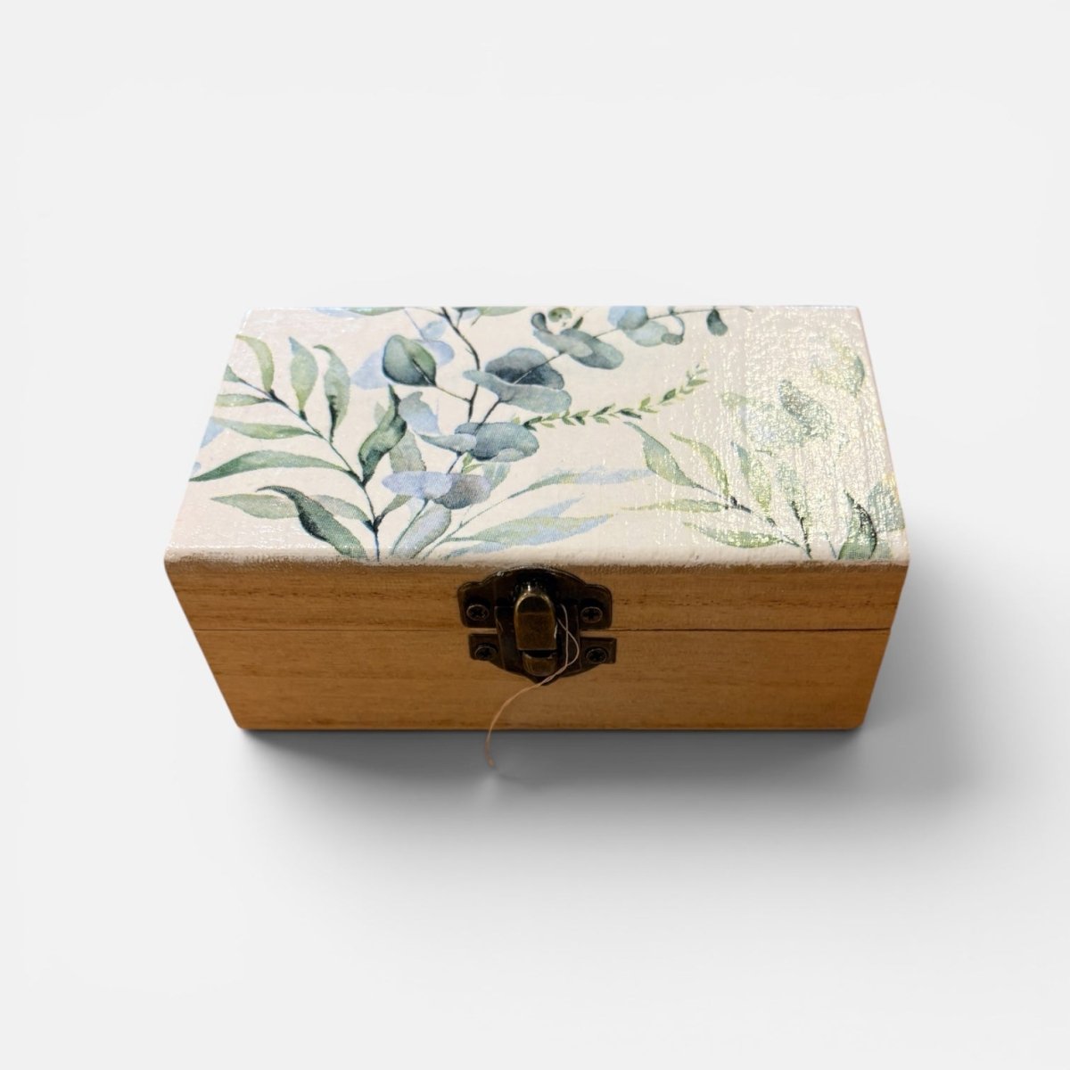 A rectangular wooden box with a light natural wood base and a lid decorated with a watercolor-style botanical pattern of green leaves and blue-gray flowers on a cream background. It features a dark metal latch closure on the front, set against a plain light gray background.