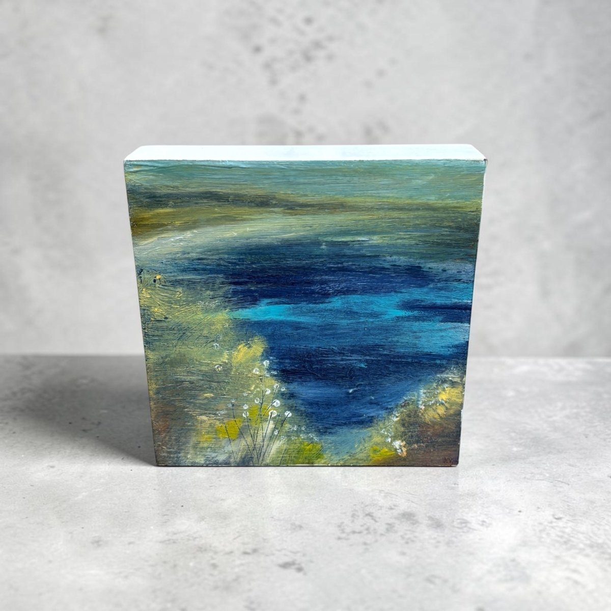 A small rectangular canvas painting of an abstract landscape with deep blue water, green and yellow foliage, and a pale sky, displayed on a light gray surface against a textured gray wall.