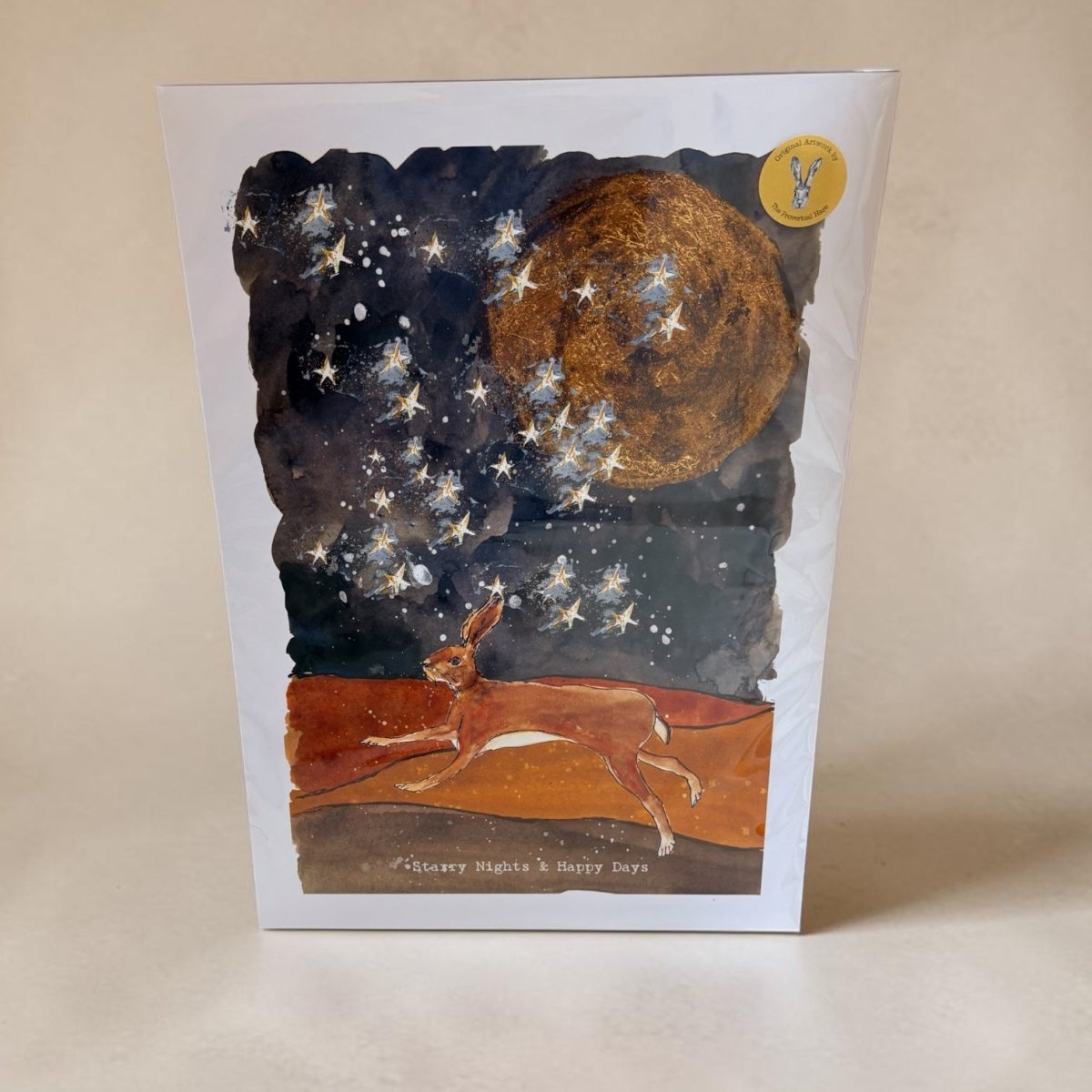 A square art print featuring a whimsical night scene with a large golden moon, scattered white stars, and a brown hare running across an orange-brown ground under a dark starry sky. The print is photographed standing upright against a plain beige background.