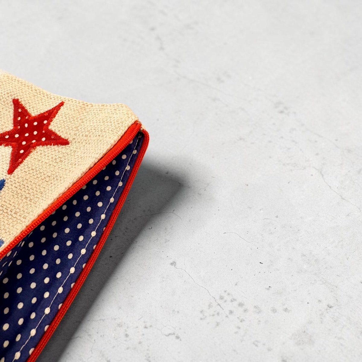 A close-up of a fabric accessory corner showing a beige woven section with a red embroidered star, a navy blue polka-dot interior, and red trim, placed on a light gray textured surface.