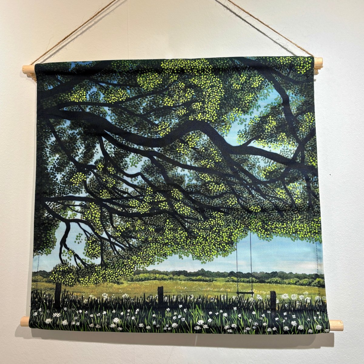 A fabric wall hanging scroll depicting a scenic landscape with a large tree, green field, flowers, and a swing, suspended by twine from wooden dowels on a white wall.