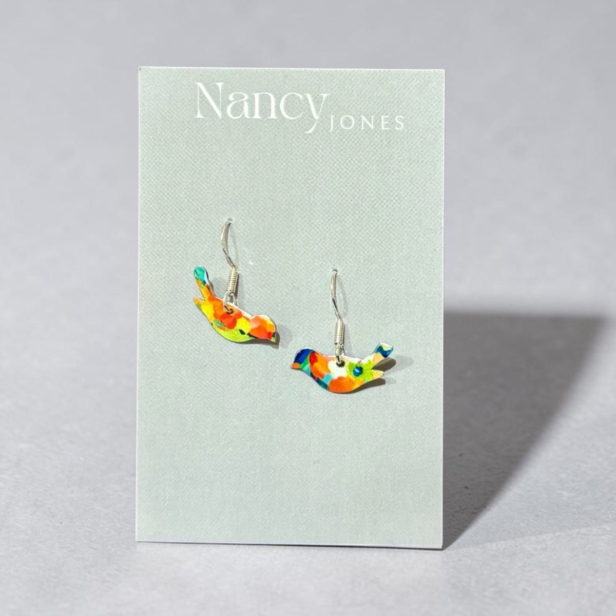 Colorful bird-shaped earrings on a Nancy Jones card against a light gray background