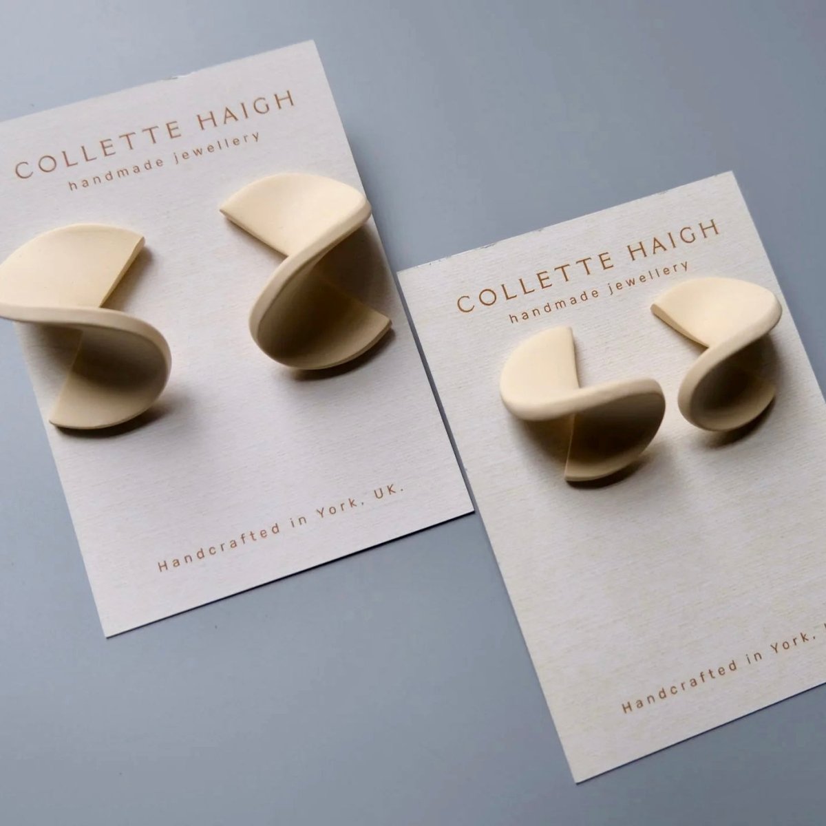 Two cream-colored spiral-shaped earrings mounted on white display cards branded Collette Haigh, photographed on a smooth light gray surface.