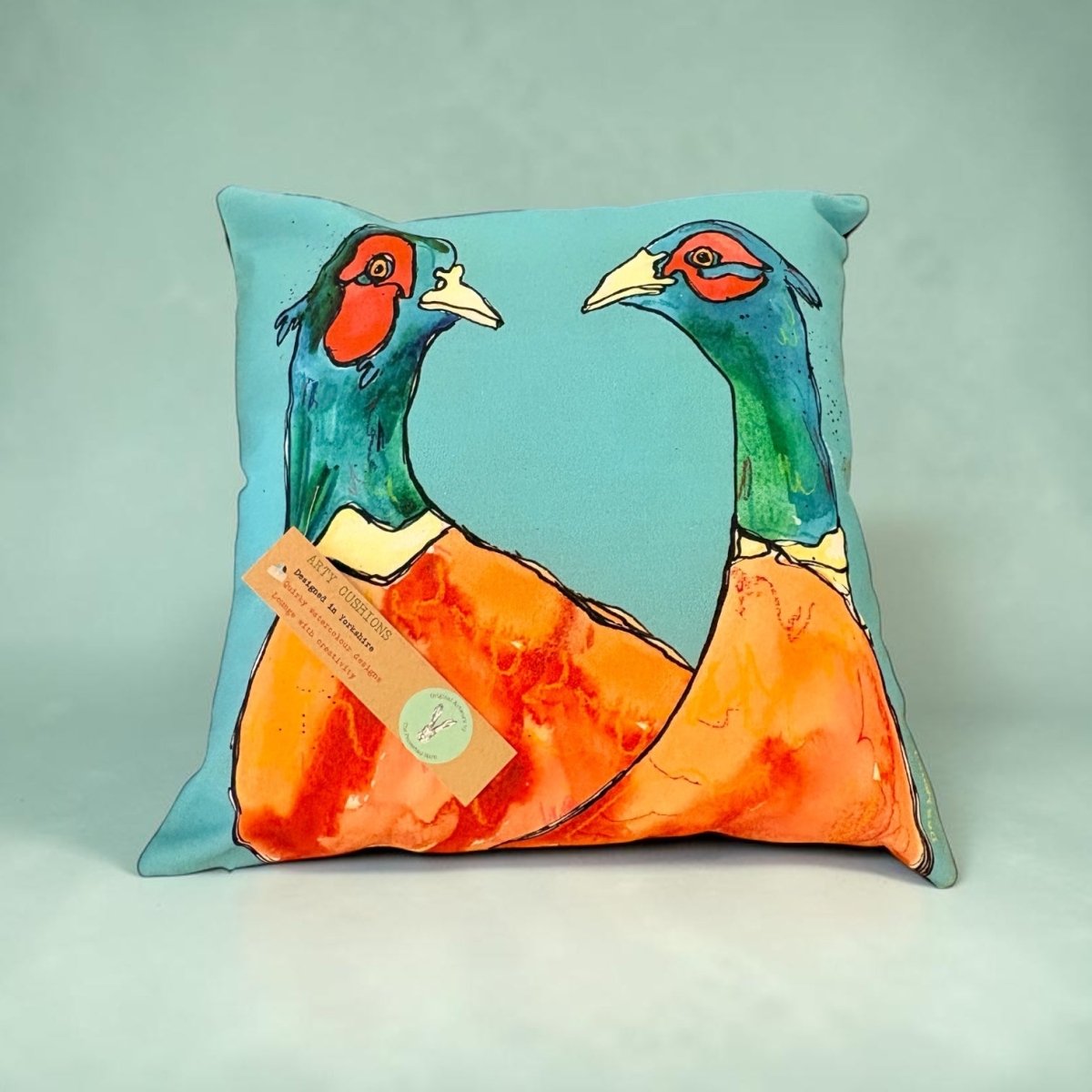 A square decorative throw pillow featuring a colorful artistic print of two pheasant birds on a light blue background, with a small paper tag attached near the lower left corner.