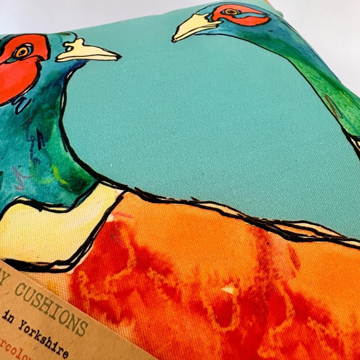 A close-up of a decorative throw pillow featuring a colorful printed design of pheasants on a turquoise background, with part of a brown paper label visible at the bottom left.