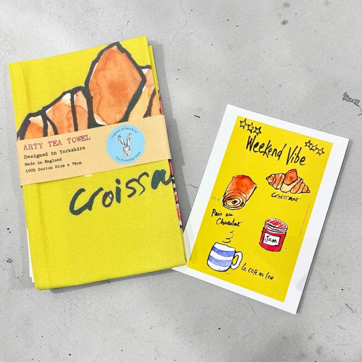 Yellow tea towel with croissant design and matching card on a gray surface